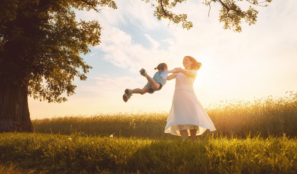 An adult wearing a white dress spins a child in the air in a grassy field at sunset, capturing a joyful moment that highlights the healing power of trauma therapy, with trees and clear sky in the background.